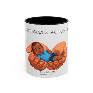 May include: A white ceramic mug with a black interior and rim. The front features a graphic of two large hands cradling a baby wearing a blue shirt and dark shorts. Above the graphic is the text GOD'S AMAZING WORK OF ART and below is Jeremiah 29:11 Jeremiah 1:5.