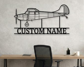 May include: A black metal wall art piece featuring the silhouette of a single-engine propeller airplane. Below the aircraft, a horizontal bar contains the text CUSTOM NAME in a bold, serif font, supported by three vertical metal rods.