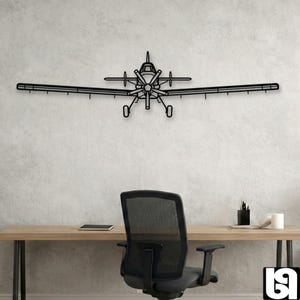 Könnte beinhalten: Schwarze Metall-Wandkunst in Form eines Flugzeugs. Das Flugzeug hat einen Propeller, Flügel und eine lange Stange, die sich von jedem Flügel erstreckt. An einer hellgrauen Wand über einem Schreibtisch und Stuhl montiert.