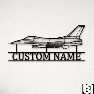 May include: Black metal wall art depicting a fighter jet outline. The design includes the text "CUSTOM NAME" below the aircraft, suitable for customisation. The artwork is mounted on a white background.