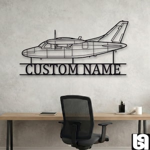 May include: A black metal wall art piece shaped like a twin-engine airplane silhouette. Below the aircraft, a horizontal bar features the text "CUSTOM NAME" in bold, capital letters. The design is mounted on a light gray textured wall.