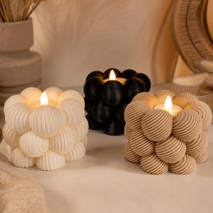 May include: Three decorative candles with a unique bubble design. One is off-white, one is black, and the third is tan. Each candle has a small, lit, faux flame in the center, creating a warm glow. The candles are arranged on a neutral surface.