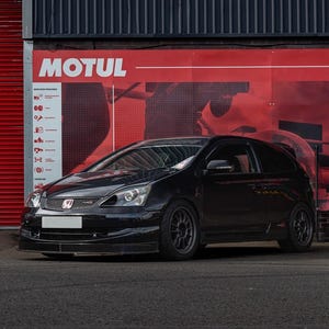 Può includere: Una Honda Civic nera con un emblema Honda rosso sul davanti. L'auto è parcheggiata davanti a un muro rosso con un logo "MOTUL" bianco.