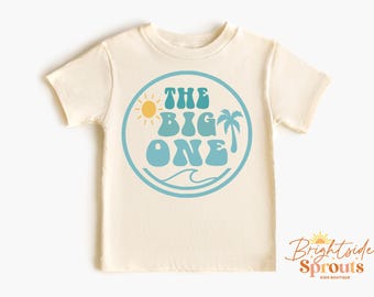May include: A light beige t-shirt with a blue and yellow graphic that says "The Big One" with a sun, palm tree, and wave design.