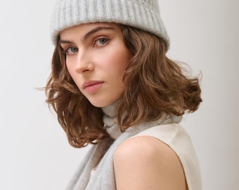 May include: A woman wearing a light gray ribbed knit beanie and matching scarf. The beanie sits close to the head, and the scarf drapes around the neck and shoulders. The woman is also wearing a sleeveless top.