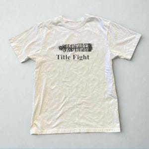 May include: Off-white t-shirt featuring a black graphic and the text "Title Fight SINCE 2003". The graphic is a stylized feather or leaf shape. The shirt has short sleeves and a crew neck.