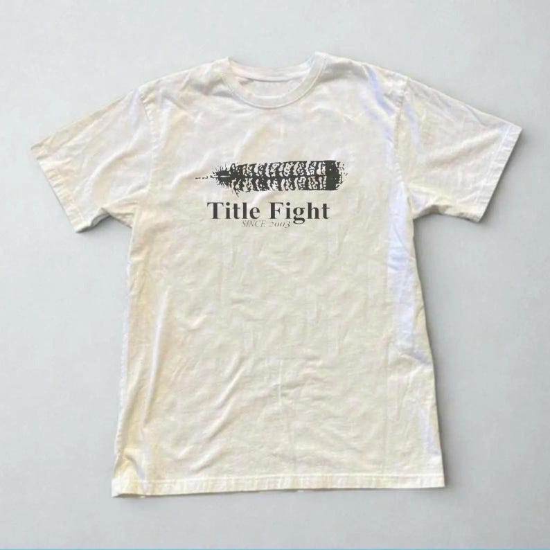 May include: Off-white t-shirt featuring a black graphic and the text "Title Fight SINCE 2003". The graphic is a stylized feather or leaf shape. The shirt has short sleeves and a crew neck.