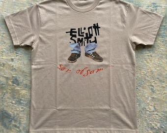 May include: A beige short-sleeve t-shirt featuring a graphic print of the text Elliott Smith in black, positioned above an image of blue jeans and brown sneakers. Below the graphic, the text son of sam is printed in a red cursive script.