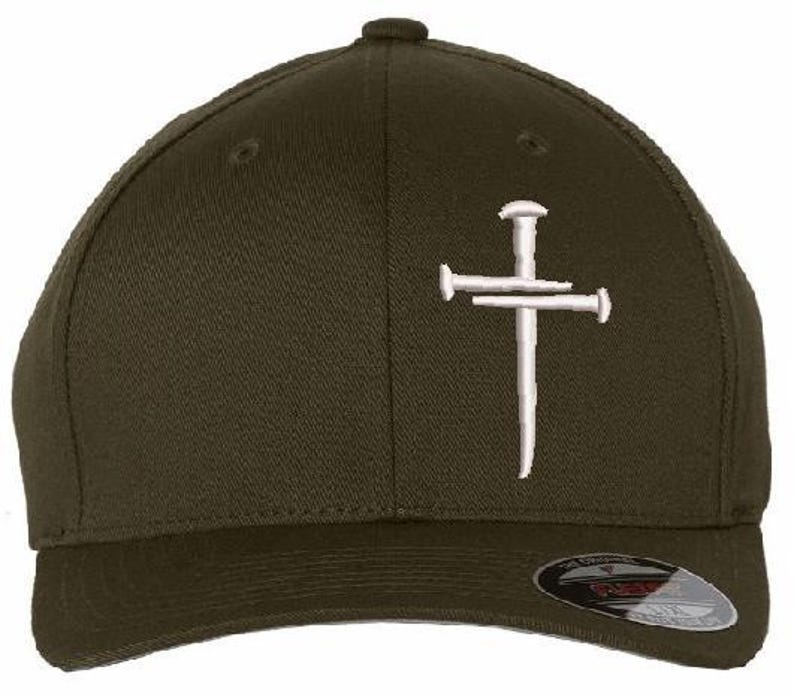 May include: Olive green baseball cap featuring a white embroidered cross made of nails. The cap has a curved brim and a small button on top. The Flexfit brand label is visible on the brim.