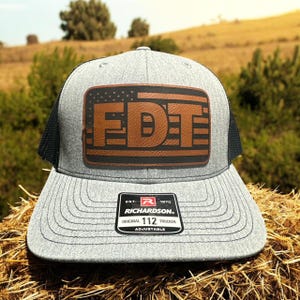May include: A gray trucker hat with a black mesh back and a brown leather patch. The patch features the letters "FDT" in a bold font, with an American flag design. The hat is adjustable.