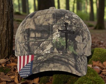 Op de afbeelding: Camouflage baseballpet met een geborduurd zwart kruis van spijkers. De pet heeft een klein Amerikaans vlagontwerp op de rand. De hoed staat tegen een bosachtergrond.