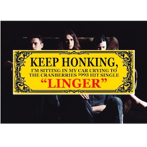May include: A yellow sign with black decorative borders and text that reads "KEEP HONKING, I'M SITTING IN MY CAR CRYING TO THE CRANBERRIES 1993 HIT SINGLE 'LINGER'". The sign is in front of a dark background with three people.