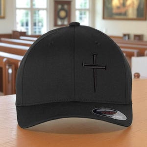 May include: A black baseball cap with a curved brim. The cap features a tonal embroidered cross design on the front. The cap is made of a textured fabric and has a fitted design.