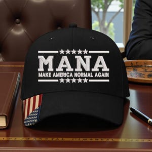 May include: Black baseball cap with the text "MANA MAKE AMERICA NORMAL AGAIN" in white. The cap features a small American flag design on the side. The cap is sitting on a wooden surface.