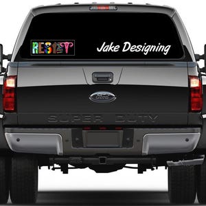 May include: Rear view of a black Ford Super Duty truck with a window decal. The decal on the top left says "RESET" in rainbow colors. The decal on the top right says "Jake Designing" in white script.