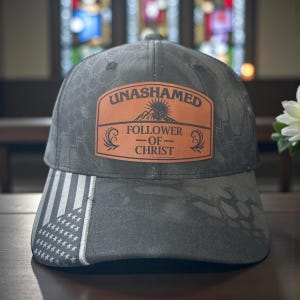 May include: A gray baseball cap with a camouflage pattern and an American flag design on the brim. The cap features a brown leather patch with the words "UNASHAMED FOLLOWER OF CHRIST" in white.