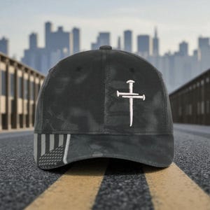 May include: Black baseball cap with a camouflage pattern and a white embroidered cross design. The cap features an American flag design on the brim. The background shows a cityscape.