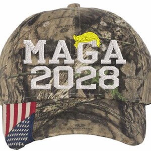 May include: A camouflage baseball cap with a white embroidered American flag on the side. The front of the cap has white embroidered text that says "MAGA 2028" and a yellow embroidered silhouette of a person's hair.
