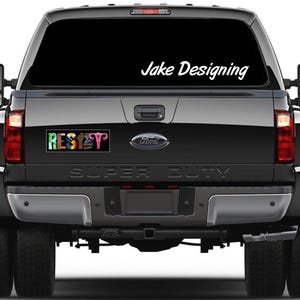 May include: A black Ford Super Duty truck with a rear window decal that says "Jake Designing" in white script. A bumper sticker below the window reads "RESET" in colorful letters, with a graphic of female reproductive organs.