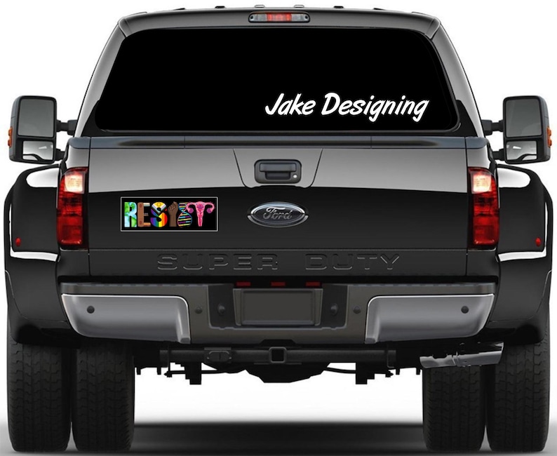 May include: A black Ford Super Duty truck with a rear window decal that says "Jake Designing" in white script. A bumper sticker below the window reads "RESET" in colorful letters, with a graphic of female reproductive organs.