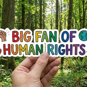 May include: A rectangular sticker featuring the text BIG FAN OF HUMAN RIGHTS in multicolored block letters. The design includes a raised fist icon, a globe, a small heart, and a scale of justice icon, all set against a white background border.