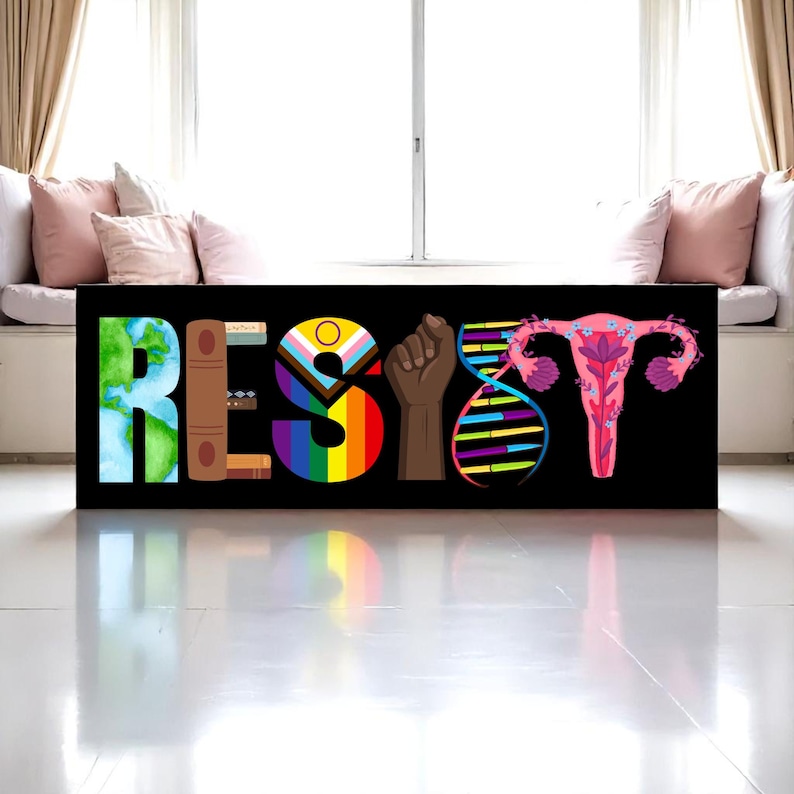 May include: A black canvas print with the word "RESIST" in colorful, symbolic imagery. The letters are composed of a world map, books, a rainbow flag, a raised fist, DNA strands, and a stylized uterus. The print is displayed in front of a window.