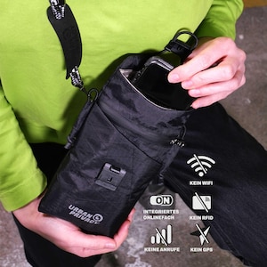 Puede incluir: Bolso bandolera negro con bolsillo con cremallera y cierre de solapa. El bolso tiene el texto "URBAN PRIVACY" impreso. Está diseñado para bloquear señales, con iconos que indican "KEIN WIFI", "KEIN RFID", "KEINE ANRUFE" y "KEIN GPS".