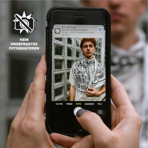 Könnte beinhalten: Ein Smartphone zeigt ein Foto einer Person mit einem Schal mit QR-Code-Muster. Der Bildschirm des Telefons zeigt eine Benachrichtigung mit dem Text "STOP TAKING PICTURES OF ME!". Das Bild enthält auch den Text "KEIN UNGEFRAGTES FOTOGRAFIEREN".