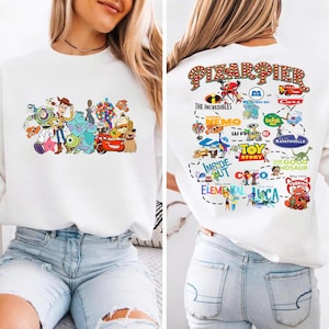 Può includere: Felpa bianca con una grafica colorata di vari personaggi Pixar sul davanti. Il retro della felpa mostra il testo "PIXAR PIER" e i loghi di diversi film Pixar, tra cui "Gli Incredibili" e "Cars".