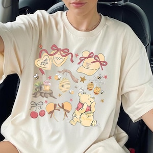 May include: Cream-colored t-shirt featuring a Winnie the Pooh design. The graphic includes illustrations of Winnie the Pooh, honey pots, bees, hearts, and the text "Winnie the Pooh". The design is in shades of red, yellow, and brown.