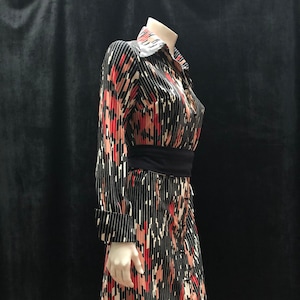 Puede incluir: Un vestido negro y blanco con un patrón geométrico rojo y rosa. El vestido tiene manga larga y cuello de camisa. Tiene un cinturón negro alrededor de la cintura.