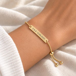 Könnte beinhalten: Ein verstellbares goldfarbenes Armband mit einer rechteckigen Metallplatte, die mit dem Text E & B 12.06.2025 graviert ist. Das Armband besteht aus einer feinen Boxkette und verfügt über einen Schiebeperlenmechanismus.
