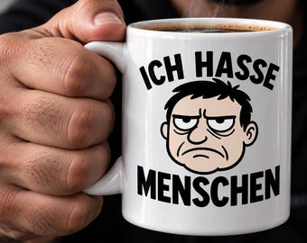 May include: A white ceramic mug featuring a cartoon illustration of a grumpy face with dark hair and the black text "ICH HASSE MENSCHEN" printed above and below the face. The mug has a curved handle and contains a dark liquid, likely coffee.
