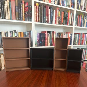 May include: A collection of miniature bookshelves in varying sizes and colors, including brown and black. The shelves are empty, and the background features a full bookshelf with numerous books. The scene is set on a dark wooden surface.