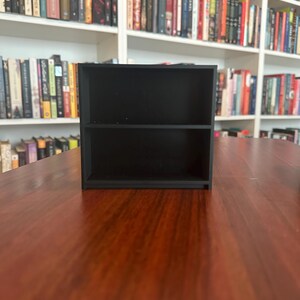 May include: A small, black, two-tiered shelf unit. The shelf is rectangular with a simple design, perfect for displaying small items. The unit is made of a solid, dark material and sits on a wooden surface. Bookshelves are visible in the background.