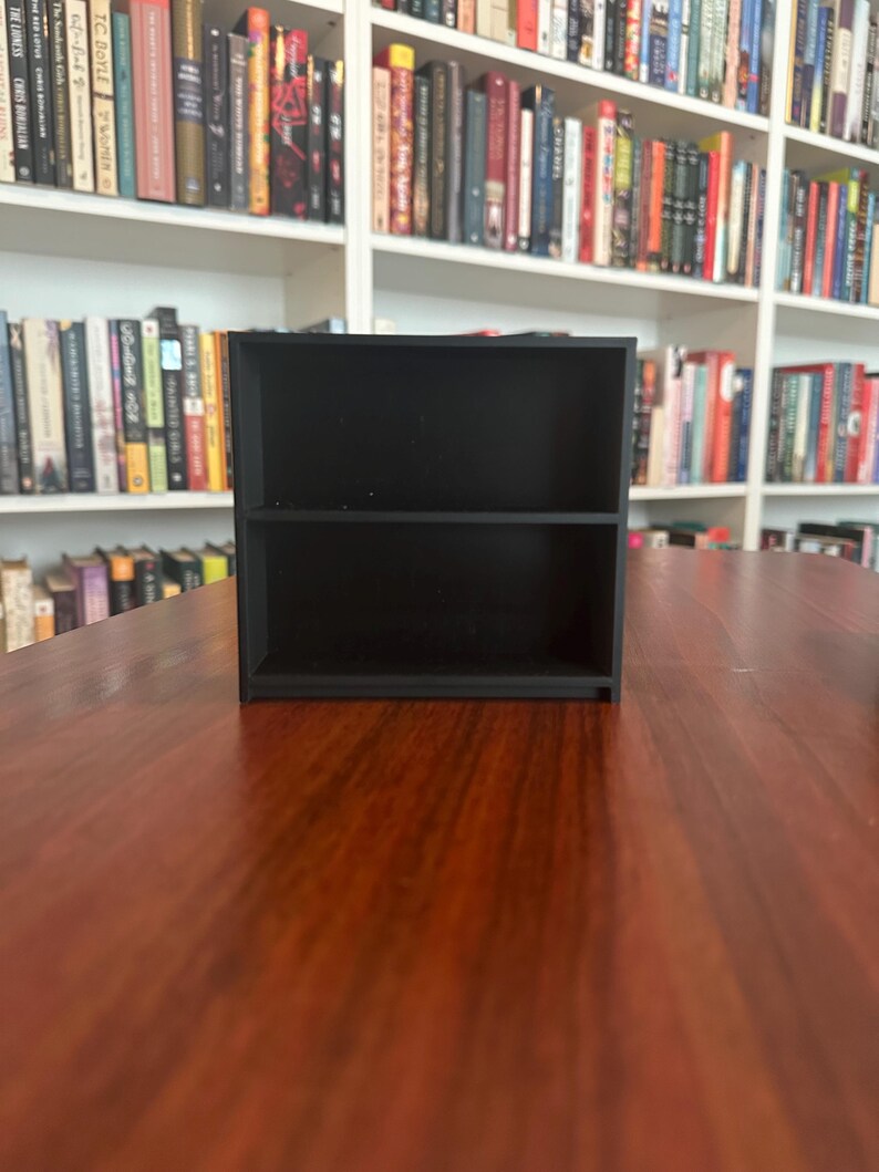 May include: A small, black, two-tiered shelf unit. The shelf is rectangular with a simple design, perfect for displaying small items. The unit is made of a solid, dark material and sits on a wooden surface. Bookshelves are visible in the background.