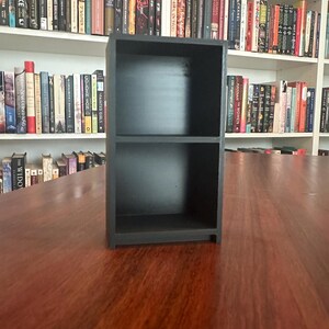 May include: A small, black, two-shelf bookcase sits on a wooden surface. The bookcase is empty, with a simple, rectangular design. Bookshelves filled with books are visible in the background.