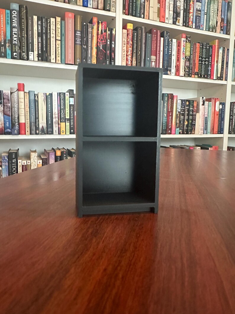May include: A small, black, two-shelf bookcase sits on a wooden surface. The bookcase is empty, with a simple, rectangular design. Bookshelves filled with books are visible in the background.
