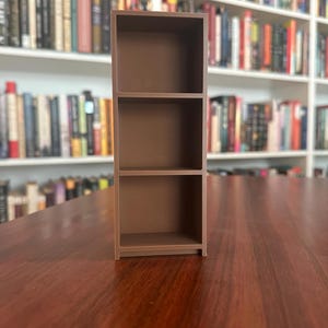 May include: A small, brown, three-shelf bookcase. The miniature bookcase is made of a solid material and is designed to hold small items. The background features a blurred image of a bookshelf filled with books.