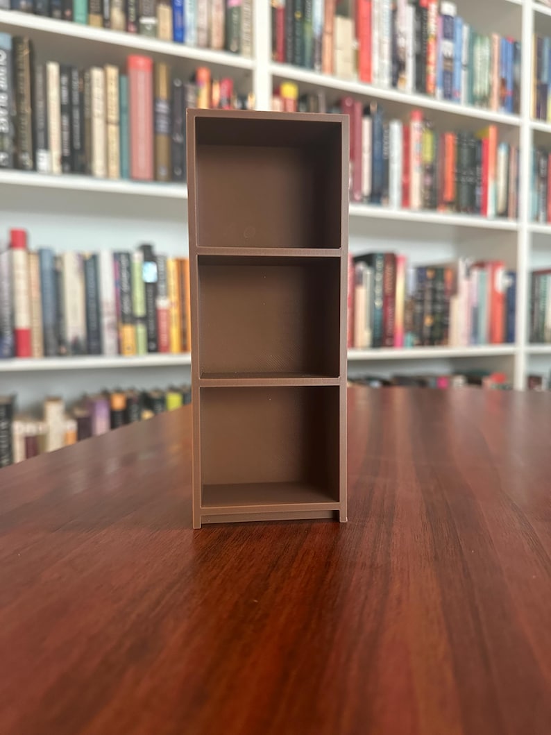 May include: A small, brown, three-shelf bookcase. The miniature bookcase is made of a solid material and is designed to hold small items. The background features a blurred image of a bookshelf filled with books.