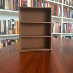 May include: A small, three-tiered brown bookshelf made of wood. The shelves are empty, and the unit is placed on a dark wooden surface. The background features a wall of bookshelves filled with books.