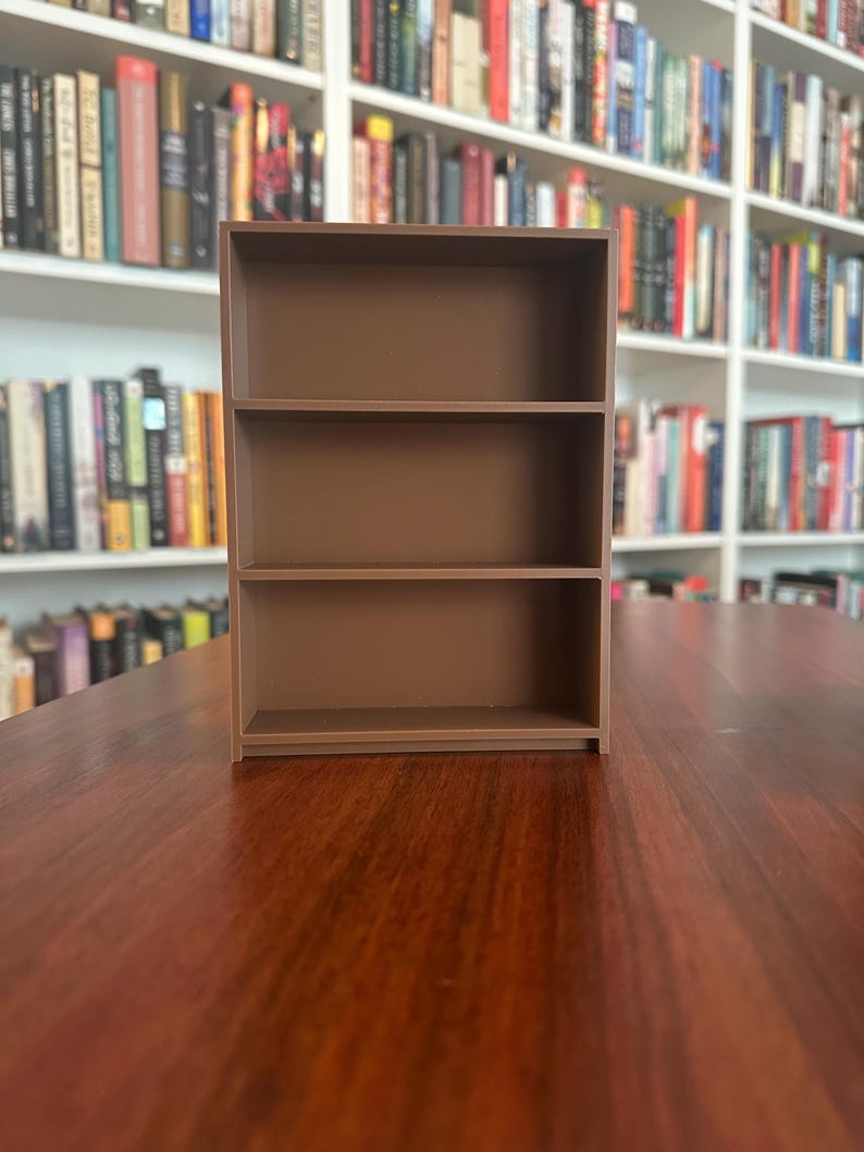 May include: A small, three-tiered brown bookshelf made of wood. The shelves are empty, and the unit is placed on a dark wooden surface. The background features a wall of bookshelves filled with books.