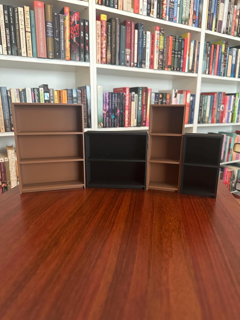 May include: A collection of miniature bookshelves in varying sizes and colors, including brown and black. The shelves are empty, and the background features a full bookshelf with numerous books. The scene is set on a dark wooden surface.