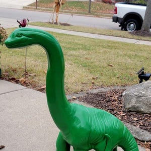 May include: A bright green dinosaur statue with a long neck and a curved tail. The outdoor lawn ornament is made of a solid material and is painted in a glossy green finish. The dinosaur is standing on a concrete surface.