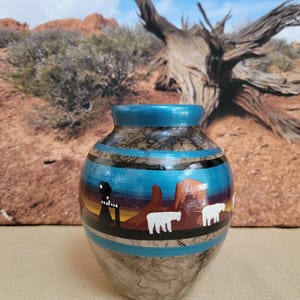May include: A small, hand-painted ceramic vase with a Southwestern desert landscape design. The vase features a blue rim, black and blue bands, and a scene with white animals and red rock formations. The base has a marbled effect.