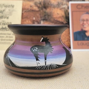 May include: Hand-painted Native American pottery vase with a horse silhouette against a sunset backdrop. The vase features a black rim and base, with a brown and purple color scheme. The design includes white accents and a unique shape.