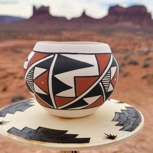 May include: A handcrafted ceramic pot with a geometric design in black, white, and red, resting on a decorative plate. The background features a desert landscape with red rock formations.