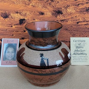 May include: A handcrafted, round, brown vase with a wide opening and a black rim. The vase features a textured surface and a painted design of figures and animals. Includes a certificate of Native American authenticity and a photo of the artist.