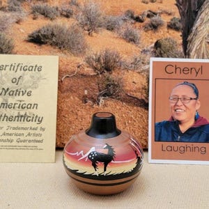 May include: A Native American pottery vase with a black and white deer design, a black top, and a certificate of authenticity. The certificate reads "Certificate of Native American Authenticity". A photo of a woman named Cheryl is also present.