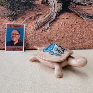 May include: A light brown turtle figurine with a blue and white design on its shell. The figurine is on a beige surface. A small photo with the text "Cheryl Laughing" is to the left of the turtle.